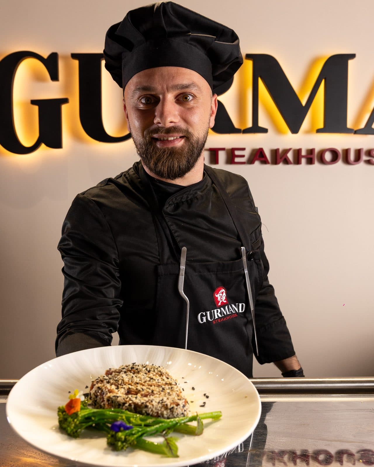 Chef prezentând un preparat în fața emblemei Gurmand